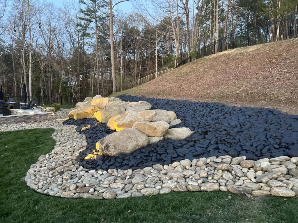 Decorative rock beds Charlotte NC style — 1400 sqft Cane River Rock installation with black stain finish by Hortus Lawn Care in Fort Mill SC